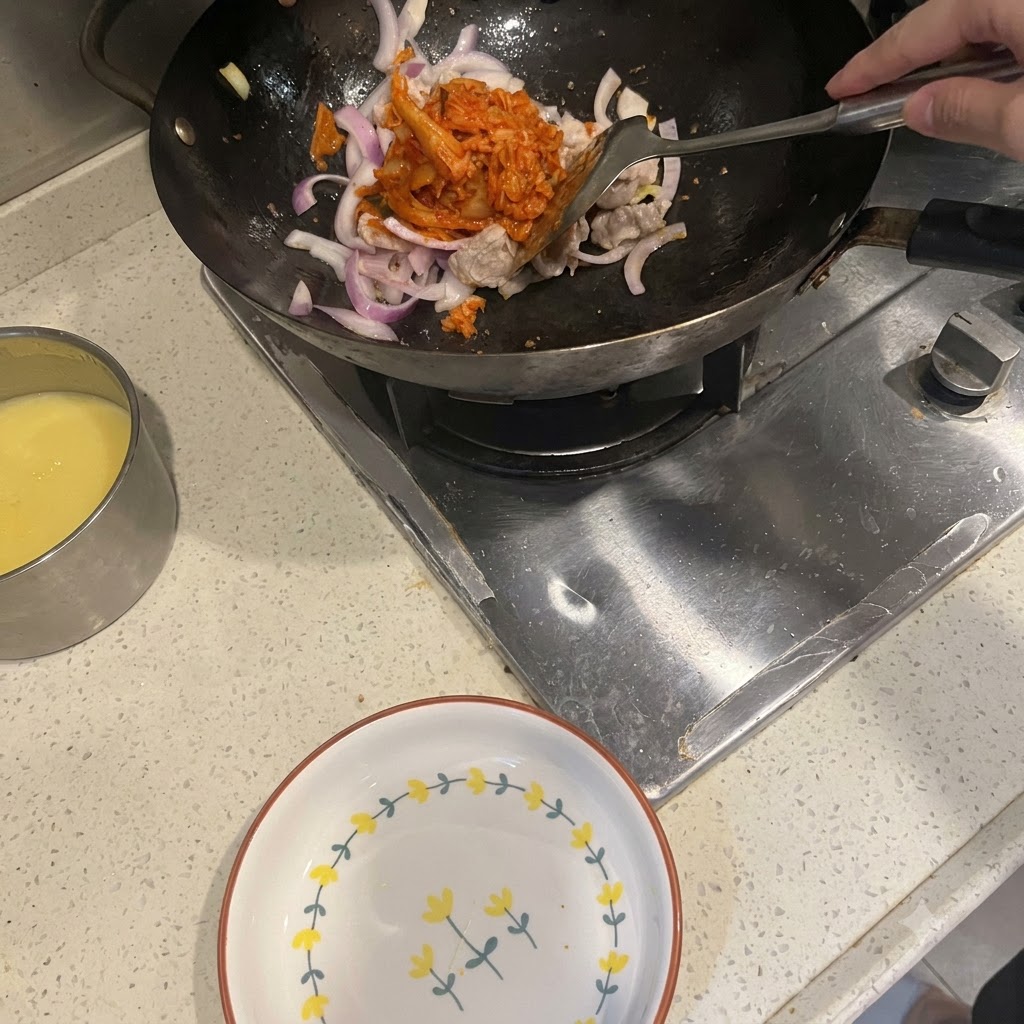 Una spatola che aggiunge kimchi rosso vivo in un wok contenente pancetta di maiale e cipolle rosse saltate.