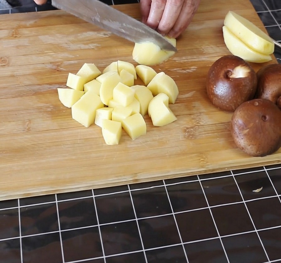 Mani che tagliano patate gialle sbucciate a piccoli cubetti su un tagliere di bambù.