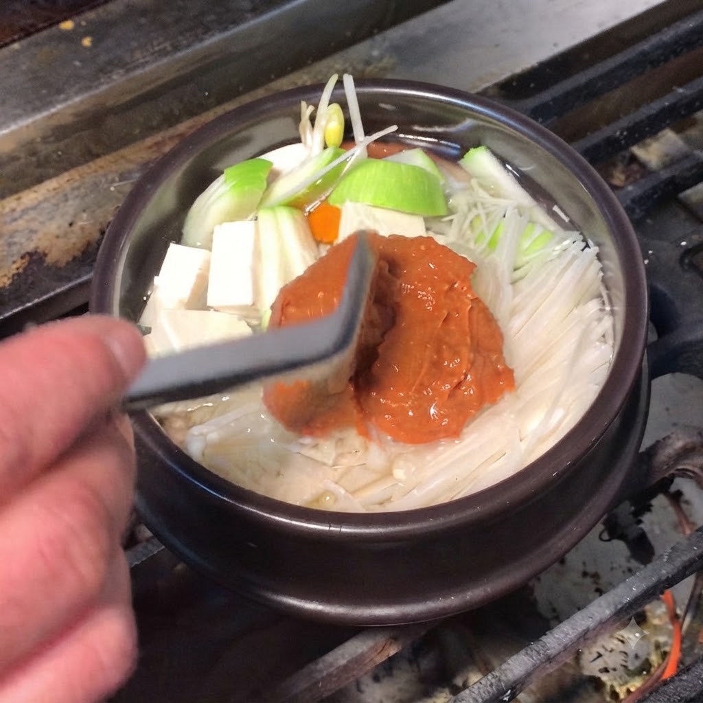 Una spatola in silicone che aggiunge ricca pasta di soia marrone e pasta di chili rossa nella pentola di cottura.