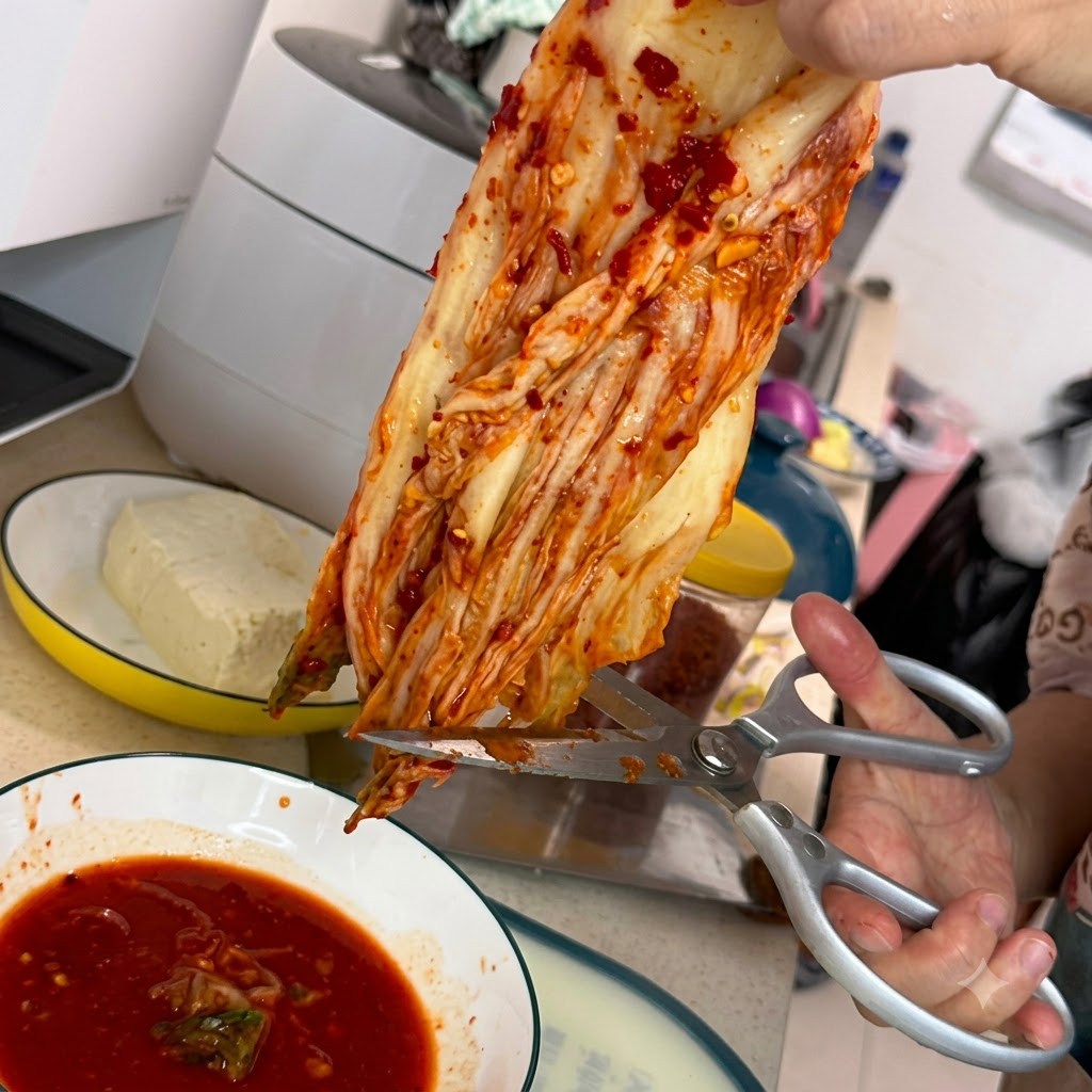 Un paio di forbici da cucina argentate che tagliano un pezzo di kimchi rosso sopra una ciotola bianca riempita di salsa rossa piccante.