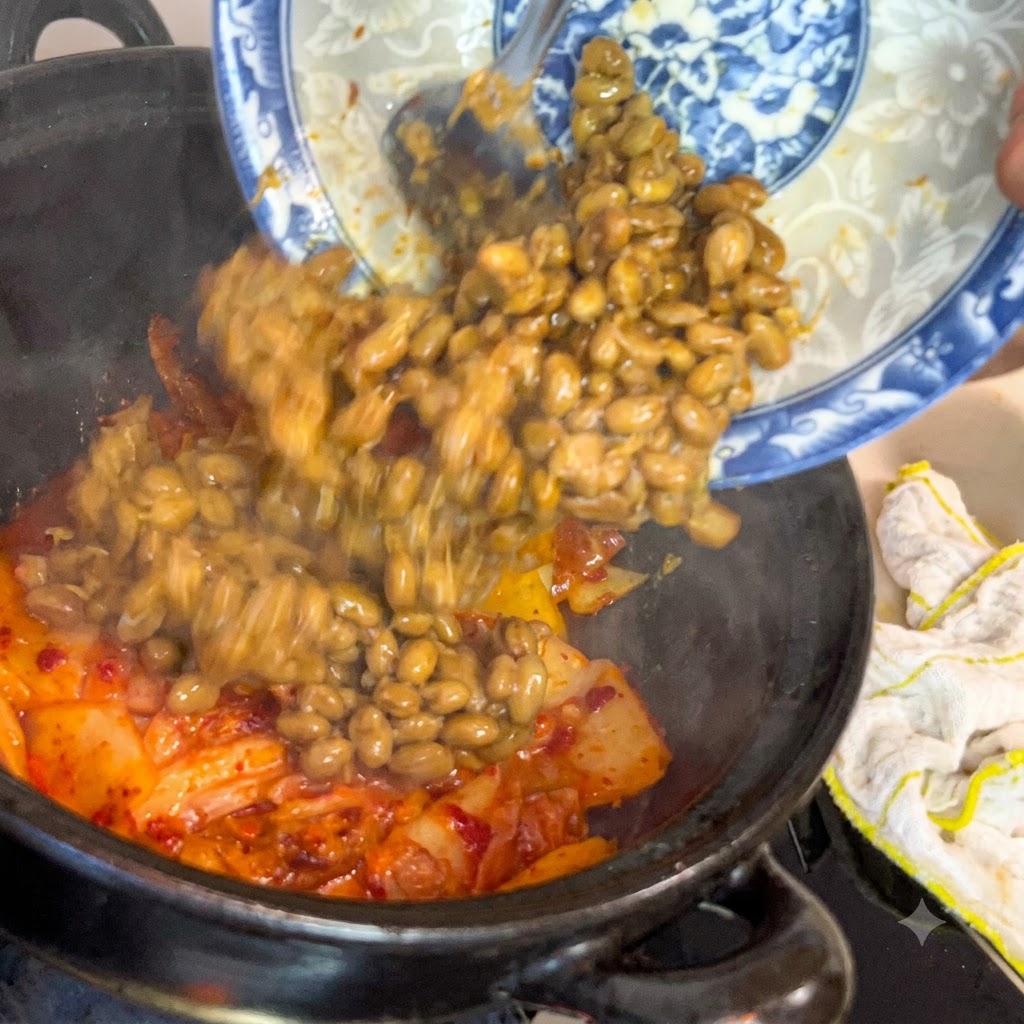 Fagioli di soia fermentati che scivolano da un piatto decorato blu e bianco in una pentola scura riempita di kimchi sfrigolante.