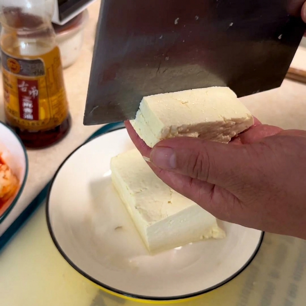 Una mano che tiene un blocco di tofu bianco mentre una mannaia lo affetta a cubetti su un piatto bianco.