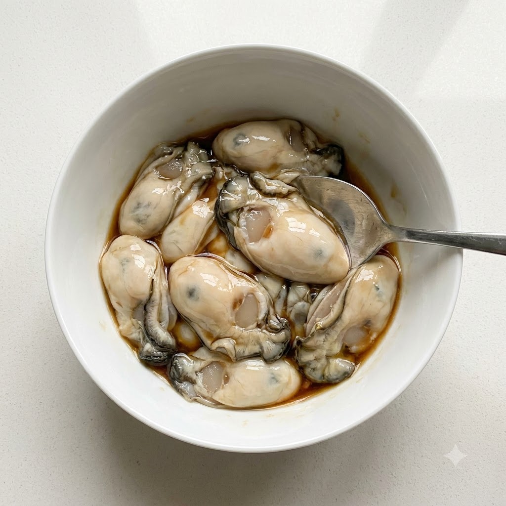 Ostriche pulite in una ciotola bianca condite con salsa di pesce e pepe bianco usando un cucchiaio.