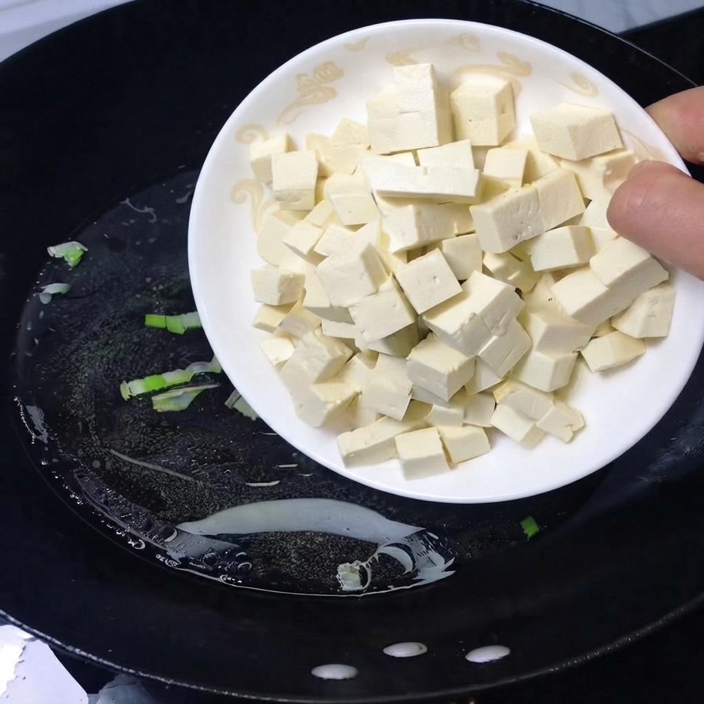 Un piatto di tofu bianco a cubetti versato in un wok nero con acqua bollente.