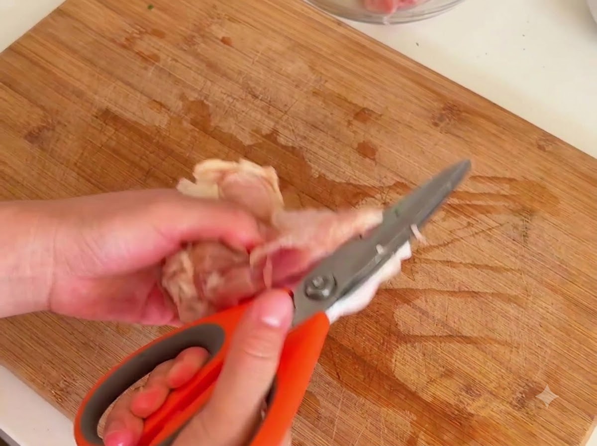 Un paio di forbici da cucina arancioni tagliano via la carne dallosso crudo di una coscia di pollo su un tagliere di legno.