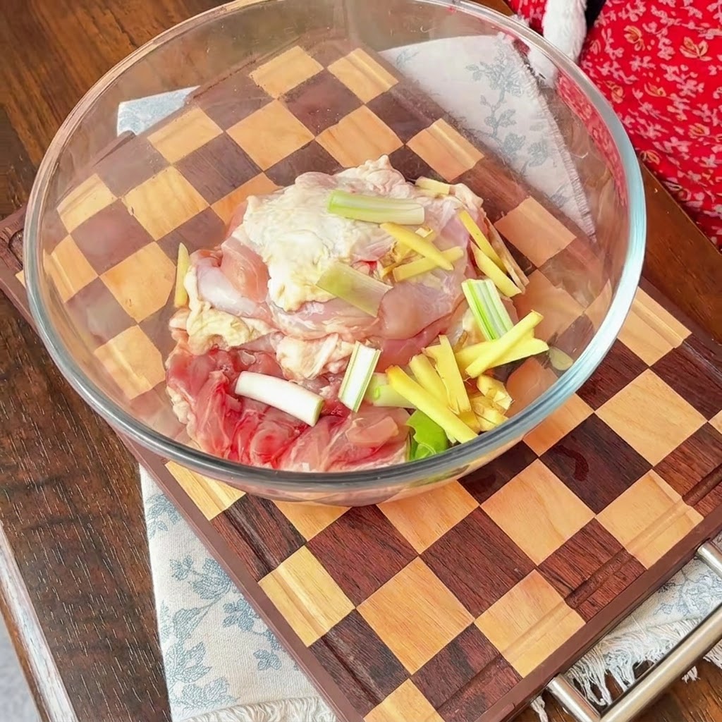 Pezzi di pollo crudo in una ciotola di vetro guarniti con cipolle rosse affettate, cipollotti e bastoncini di zenzero.