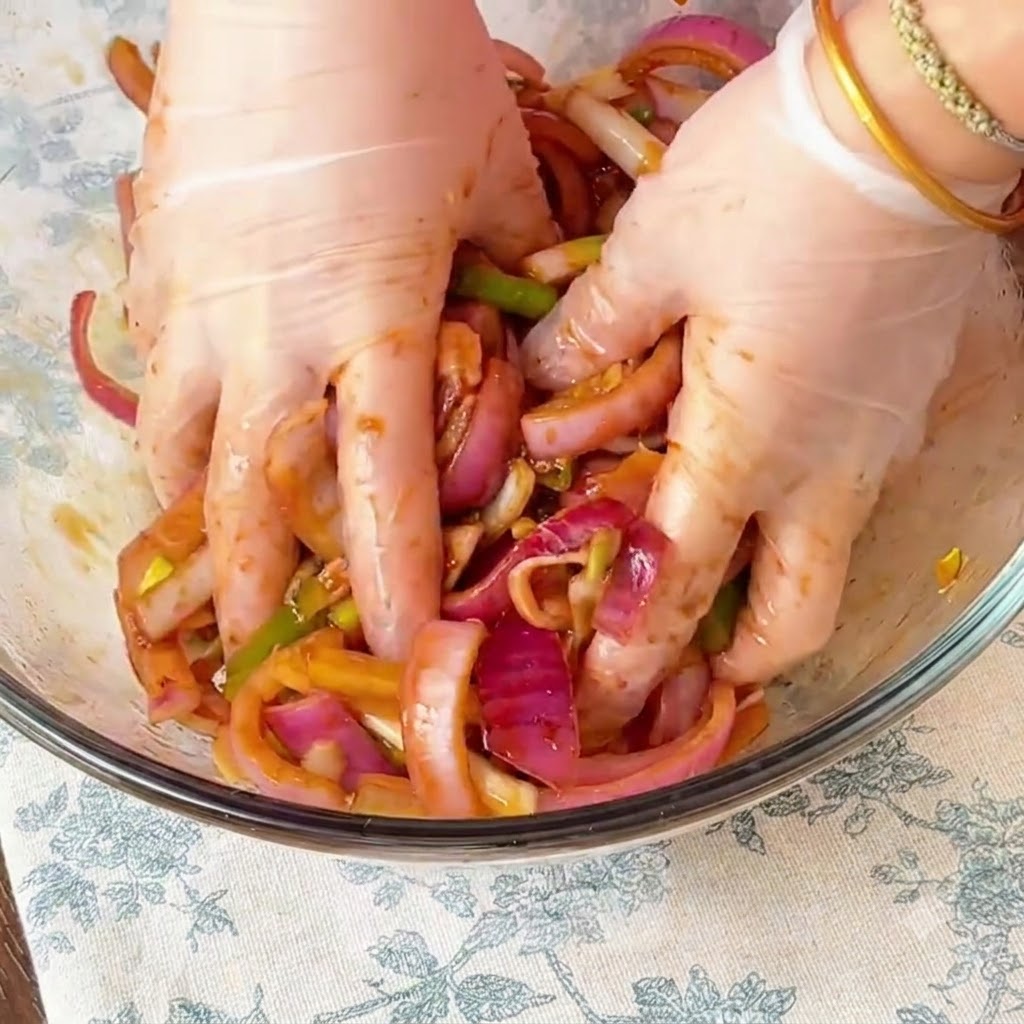 Mani con guanti che massaggiano accuratamente la marinata e le cipolle rosse affettate nel pollo allinterno di una ciotola di vetro.