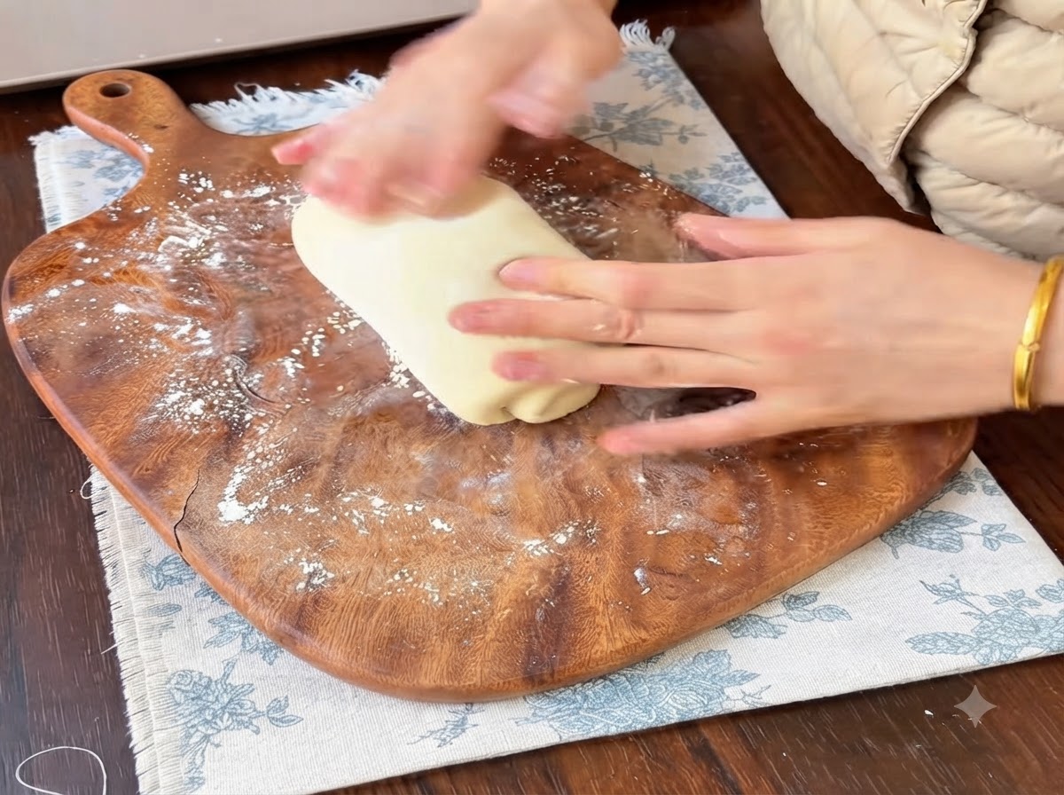 Mani che arrotolano e modellano una porzione di pasta da pane morbida su un tagliere di legno infarinato.