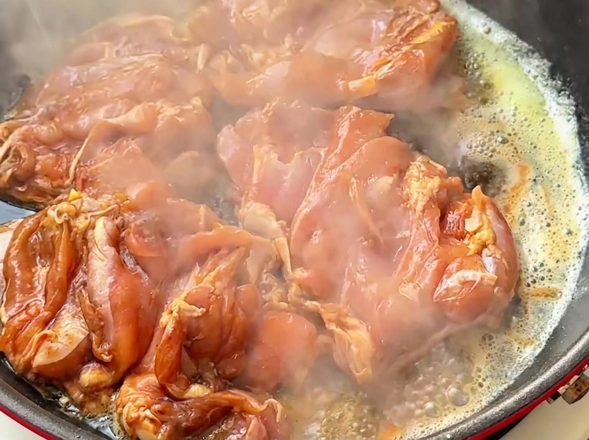 Pezzi di coscia di pollo cruda marinata che sfrigolano in una padella calda con burro e vapore che sale.