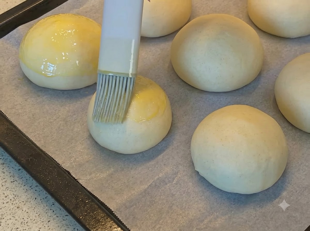 Un pennello da cucina che distribuisce luovo sbattuto dorato sulle palline di impasto lisce disposte su carta da forno.