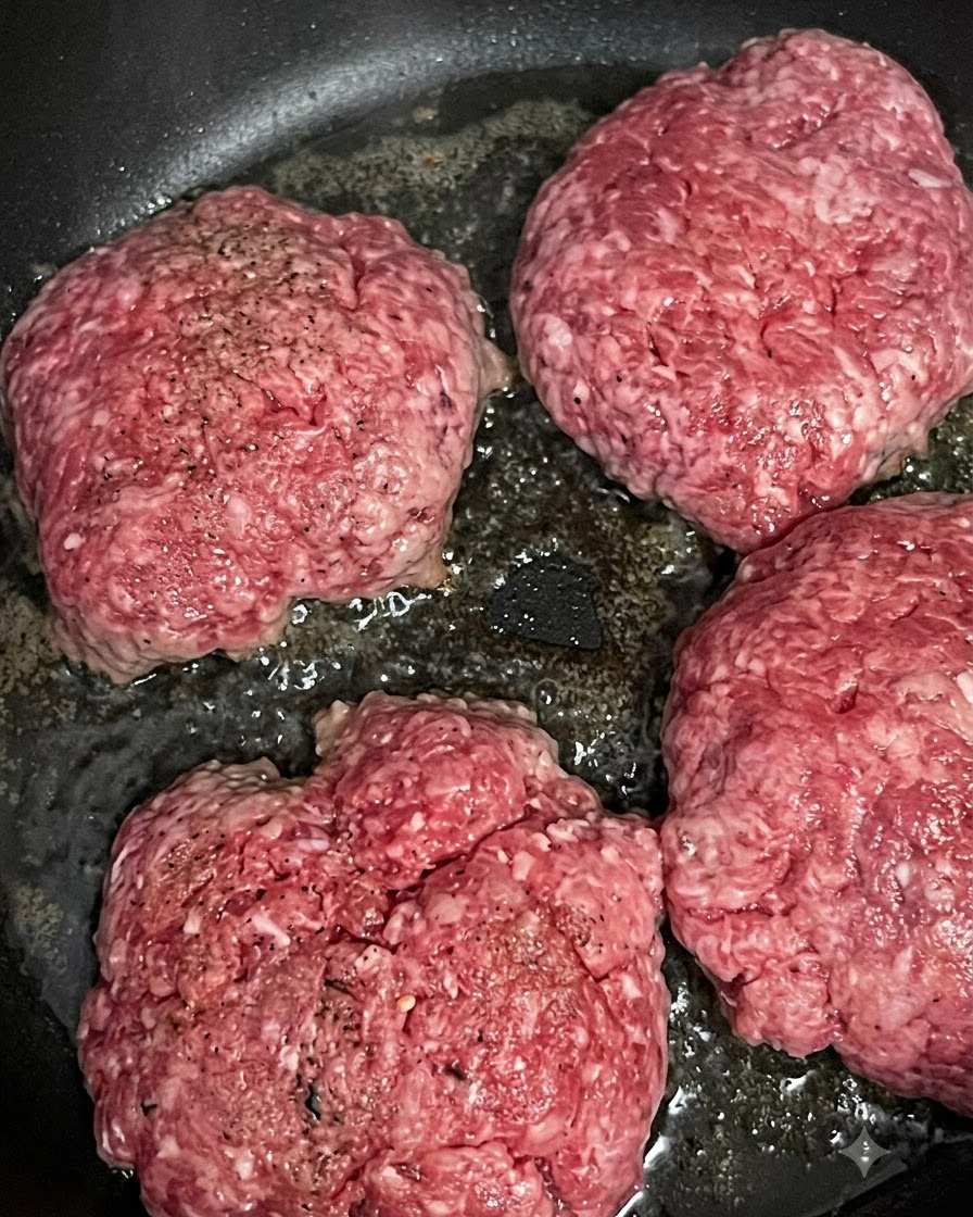 Quattro patty di manzo crudi che sfrigolano e dorano in una padella calda.