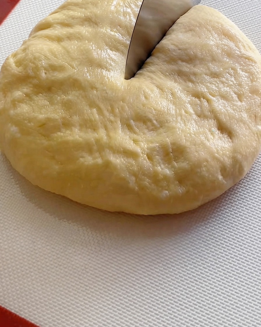 Un tagliapasta che affetta una grande palla di impasto fermentato in nove porzioni su un tappetino bianco.