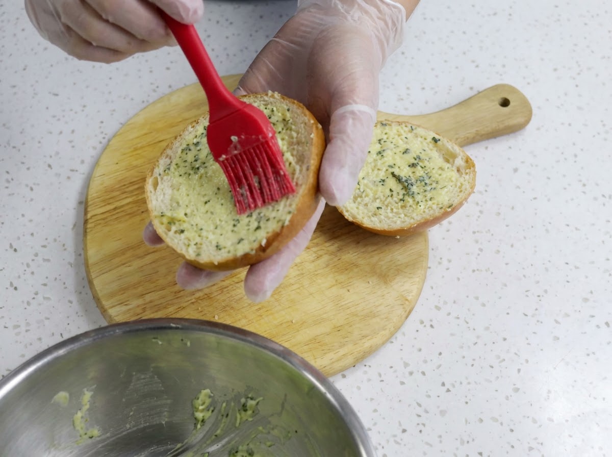 Una mano guantata che utilizza un pennello in silicone rosso per spalmare uniformemente il burro daglio sulla superficie tagliata di una metà di un panino per hamburger su un tagliere di legno.