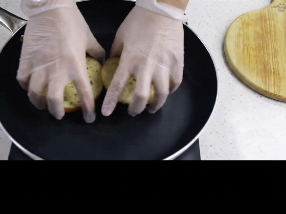 Mani guantate che posizionano il lato imburrato dei panini per hamburger rivolto verso il basso in una padella nera calda.