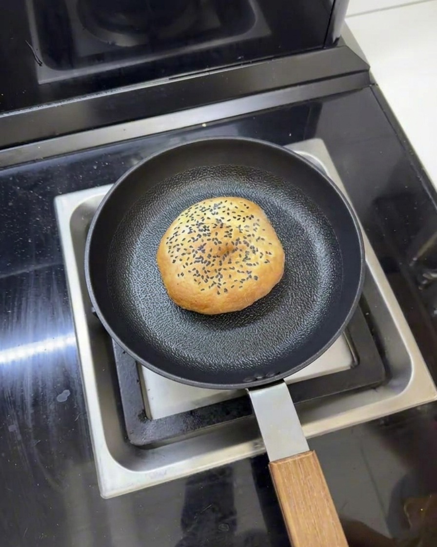 Un panino per burger al burro in una padella con acqua intorno, pronto per essere scaldato a vapore con un coperchio.