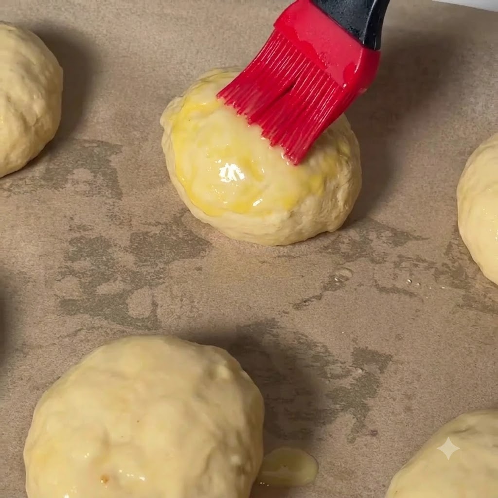 Un pennello da pasticceria rosso che applica luovo sbattuto sulla parte superiore di una palla di pasta per cheeseburger su carta da forno.