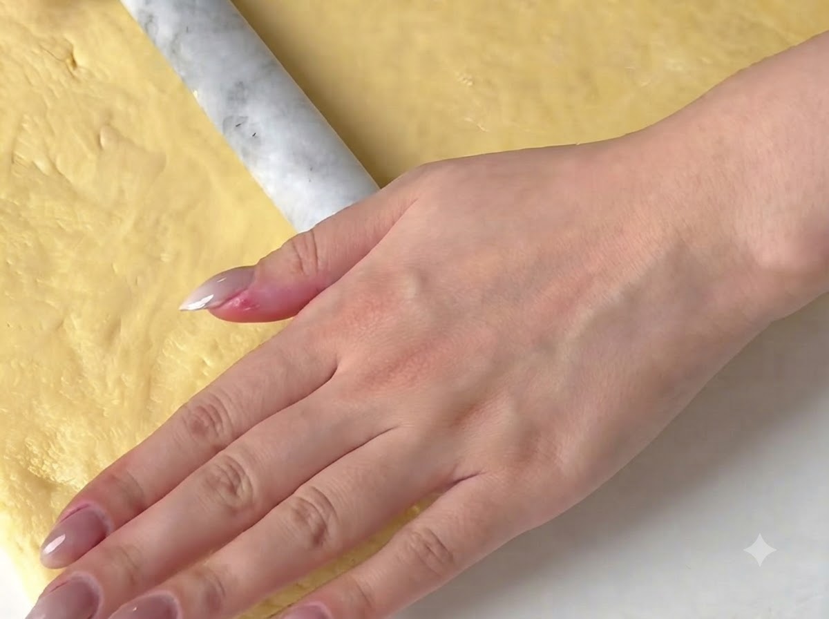 Una mano che usa un mattarello di marmo per appiattire un foglio di impasto giallo su una superficie.