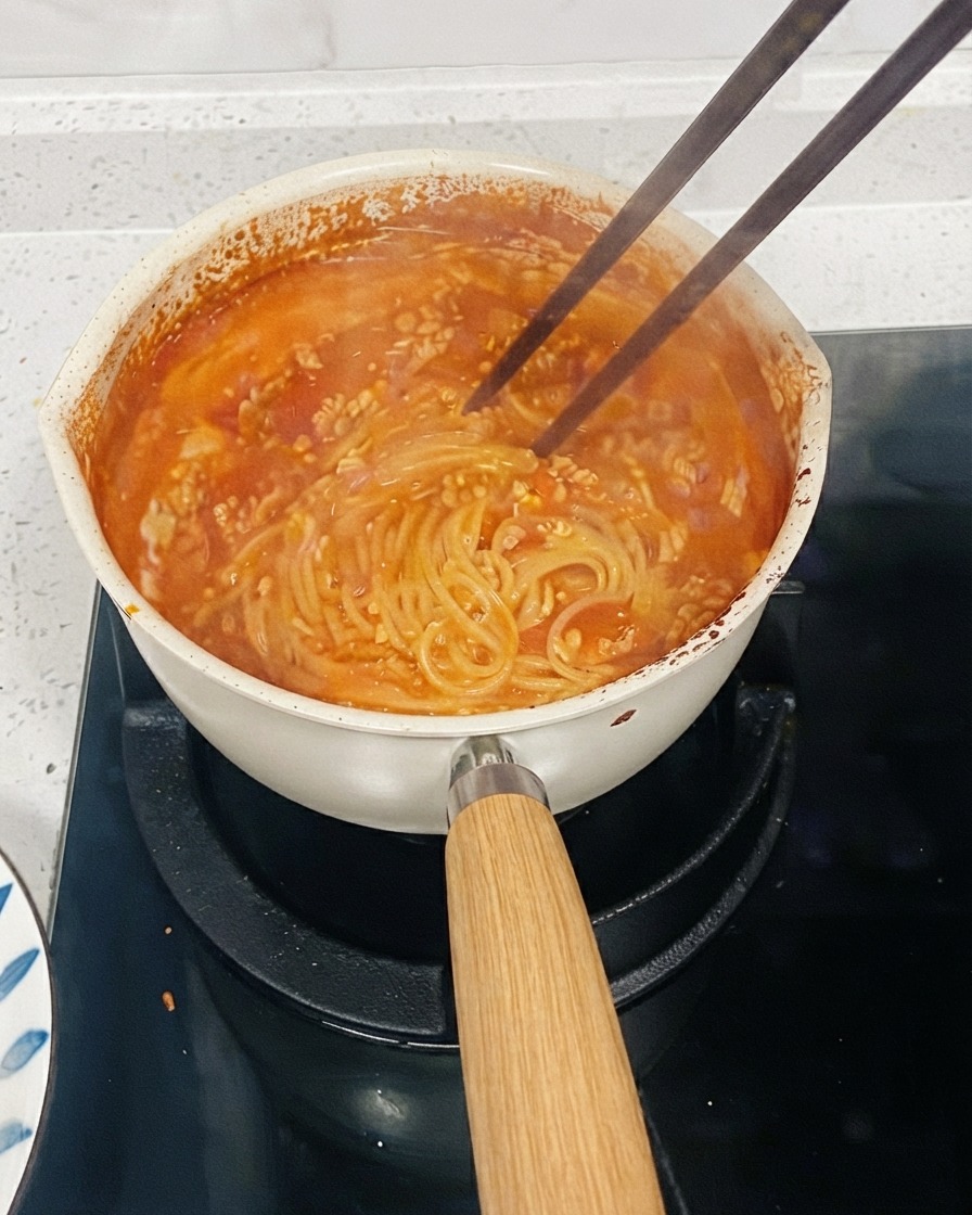 Una pentola di pasta a base di pomodoro che sobbolle su un fuoco con delle bacchette che mescolano i noodles per addensare la salsa.