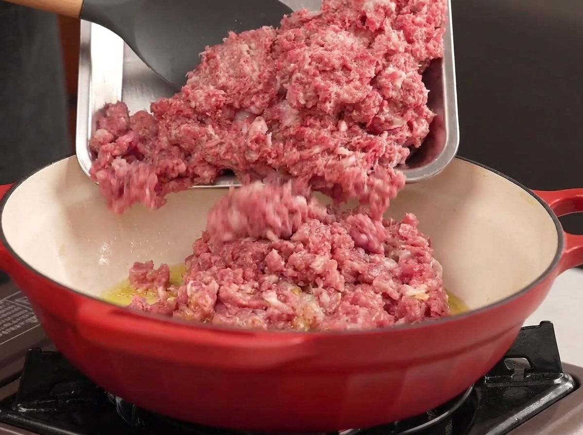 Carne macinata di manzo e maiale versata da un vassoio di metallo in una pentola rossa con olio caldo.