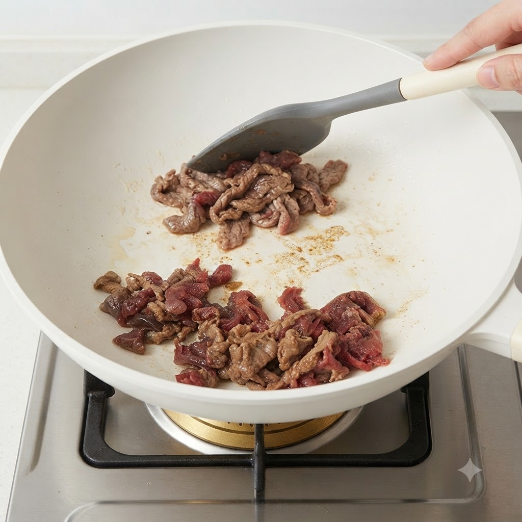 Fette di manzo marinate fatte saltare in una padella bianca usando una spatola in silicone grigia.