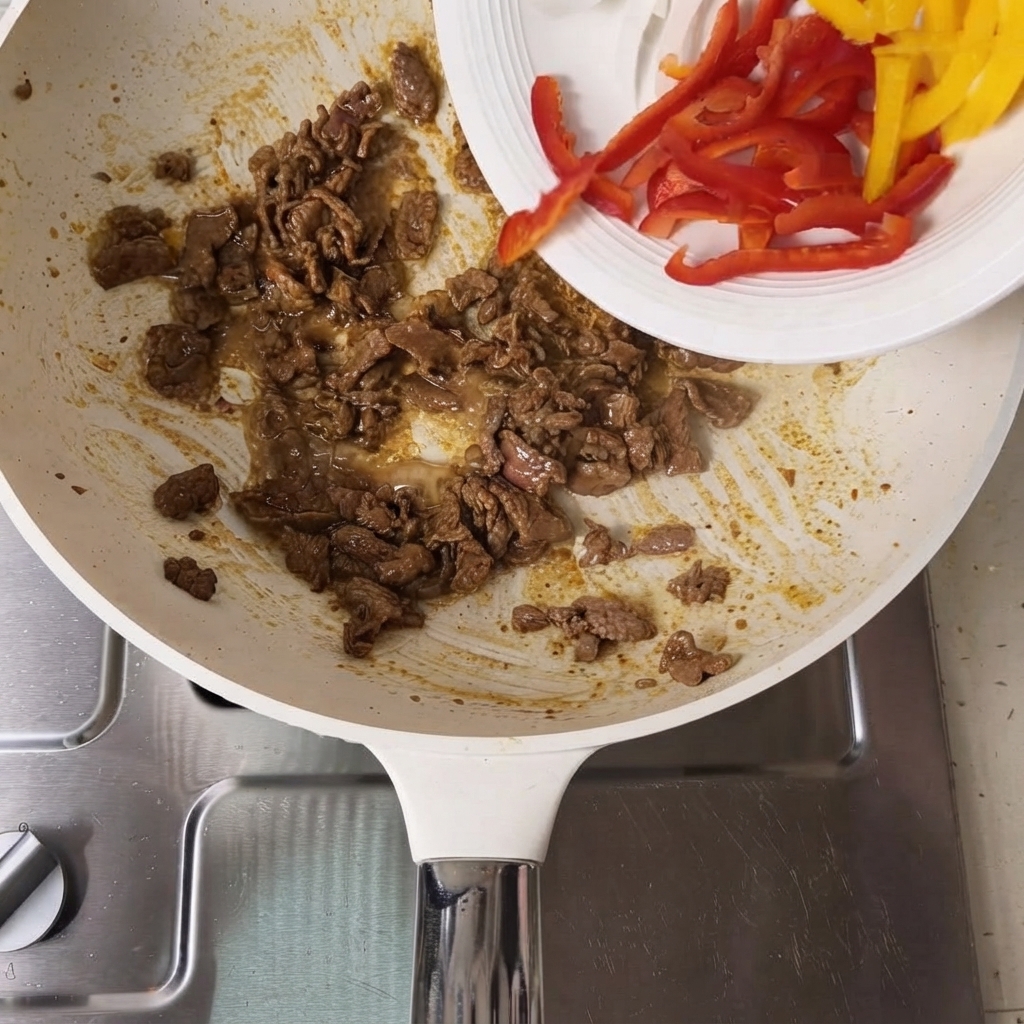 Peperoni rossi e gialli affettati versati da un piatto bianco in una padella contenente le fette di manzo cotte.