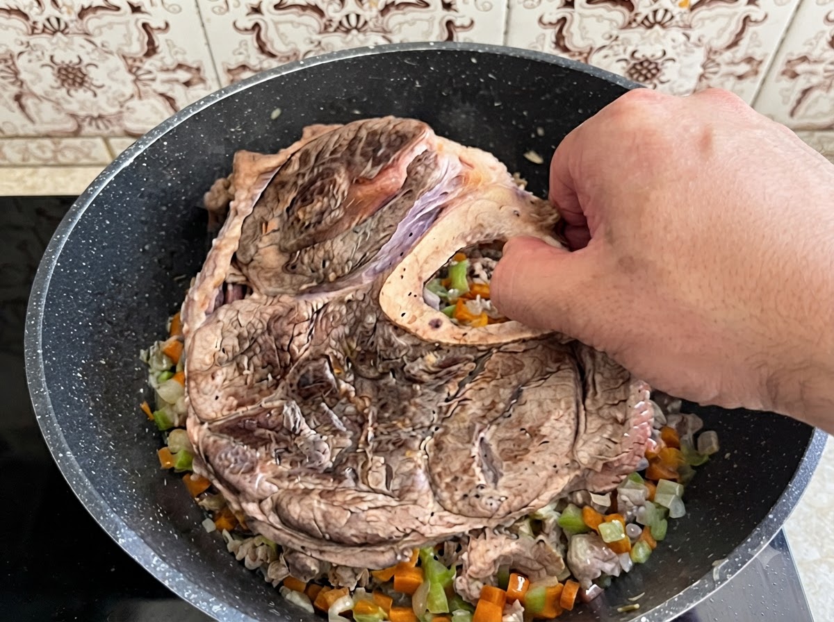 Una mano che posiziona un grosso pezzo di stinco di manzo rosolato e osso in una pentola piena di verdure cotte a dadini.