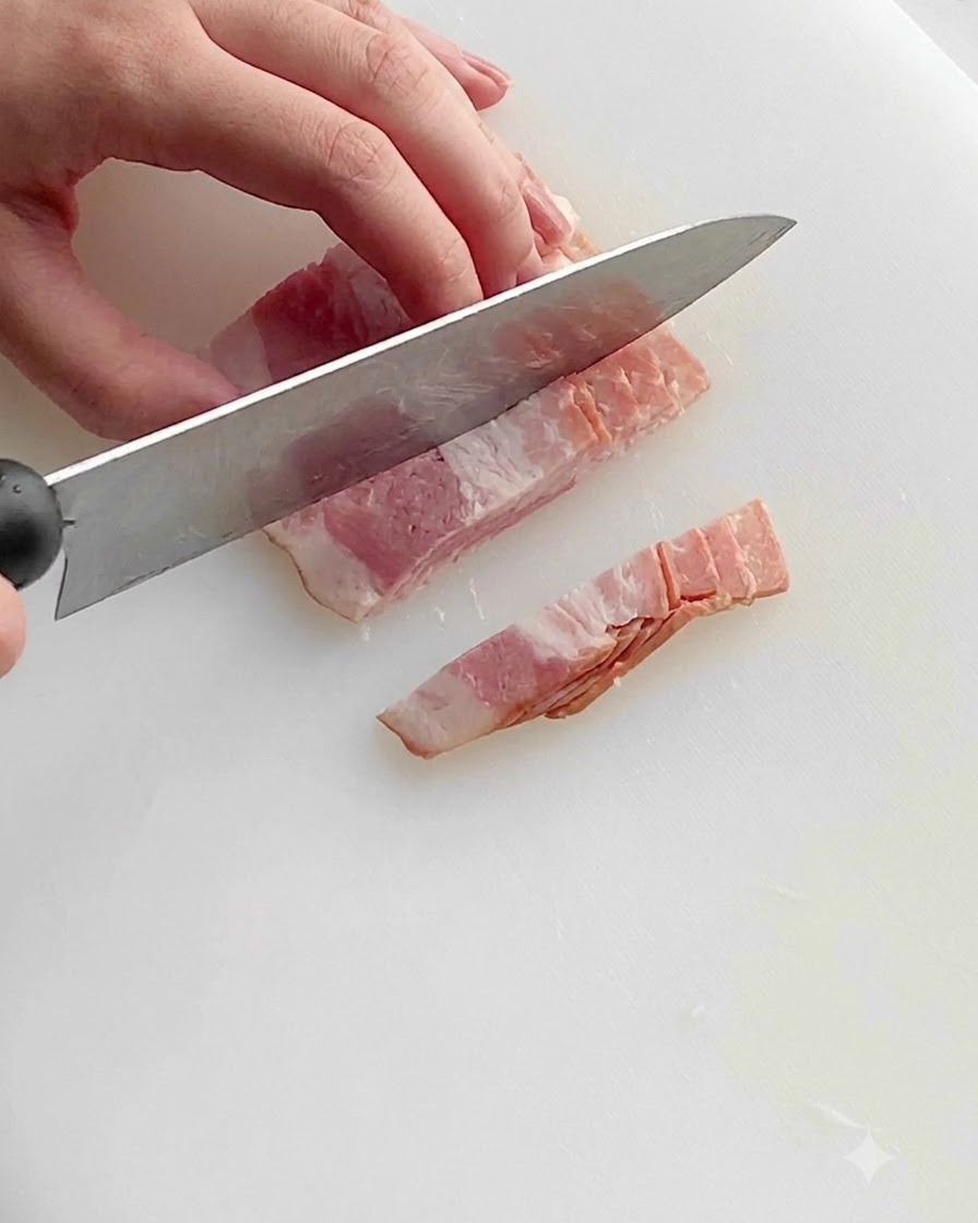 Un coltello da cucina che taglia strisce di pancetta cruda in piccoli pezzi uniformi su un tagliere di plastica bianca.