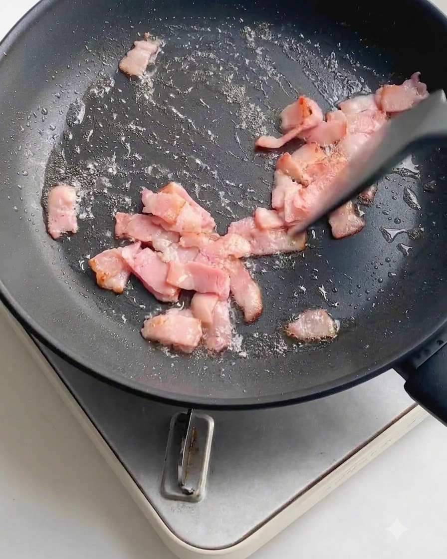 Aglio fresco tritato sparso sui pezzi di pancetta in frittura allinterno di una padella antiaderente scura.