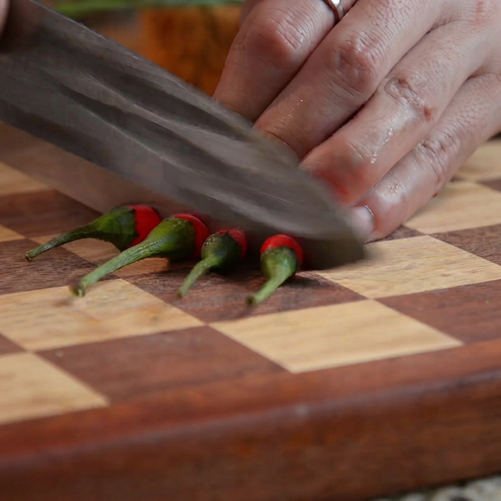 Un coltello da cucina che taglia peperoncini rossi in piccoli segmenti su un tagliere di legno.