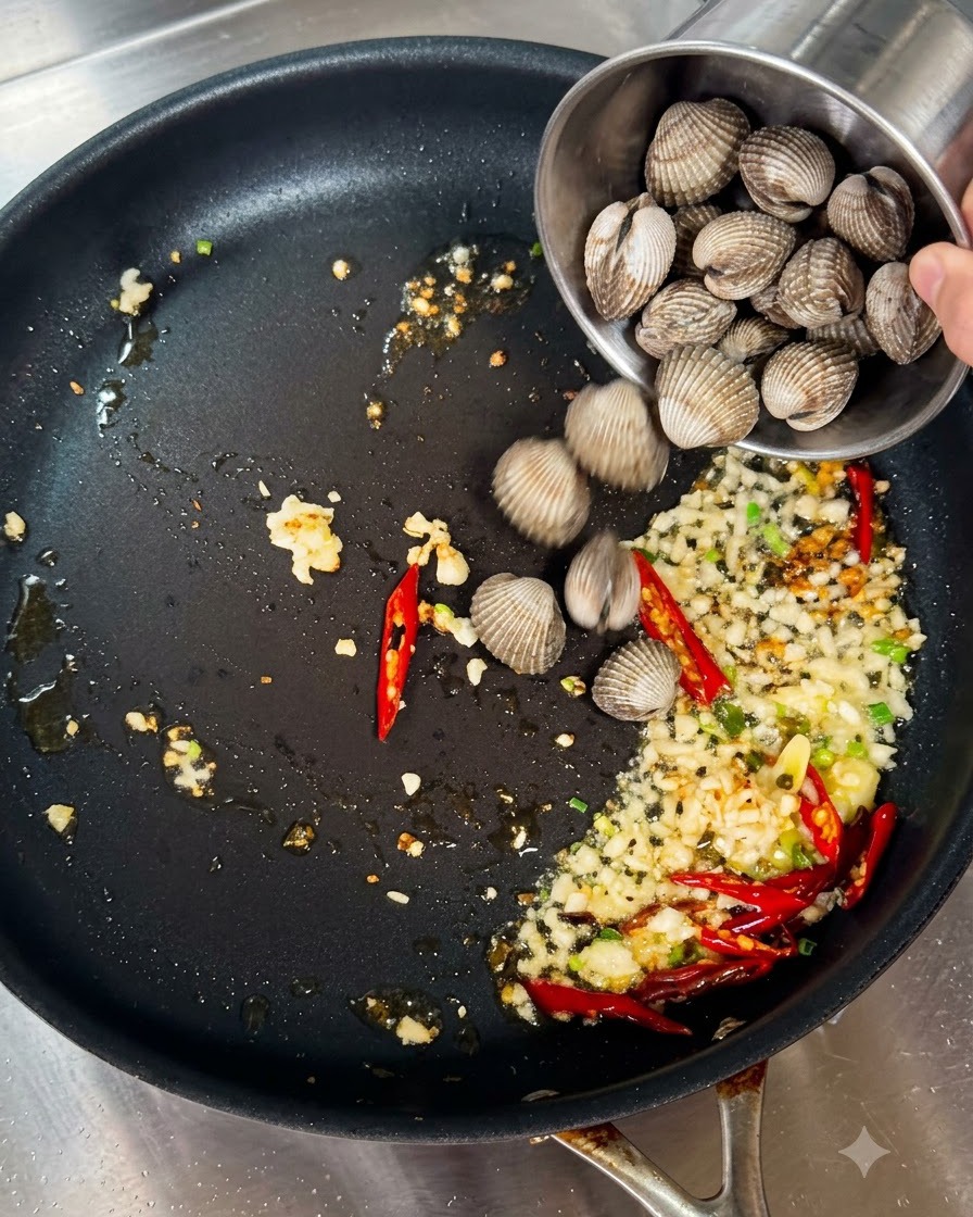 Vongole fresche versate da una piccola tazza di metallo in una padella con aglio e peperoncini saltati.