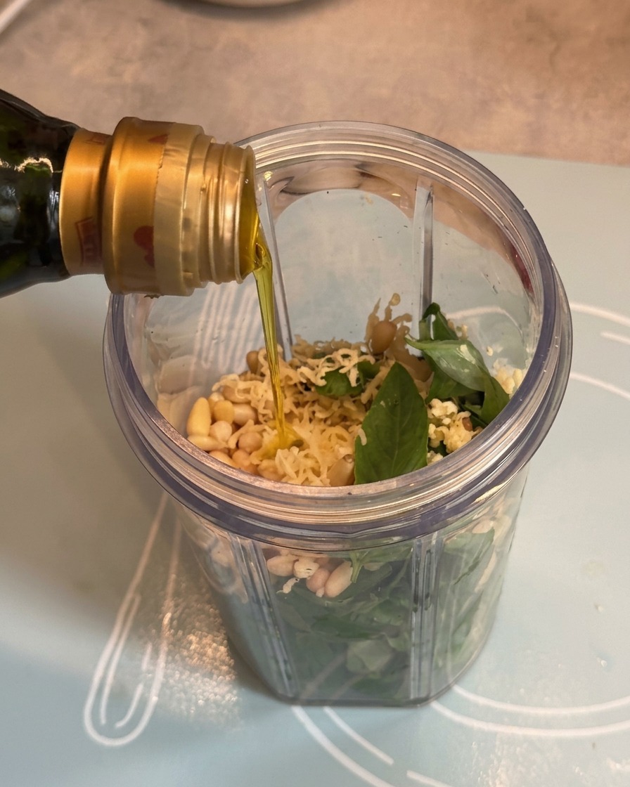 Olio doliva versato da una bottiglia nel frullatore sopra gli ingredienti del pesto.