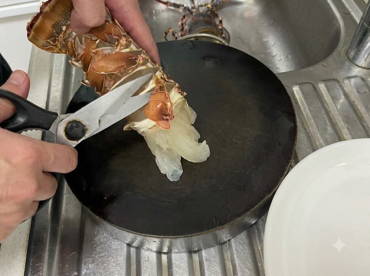 Mani che usano le forbici da cucina per tagliare lungo la membrana inferiore di una coda di aragosta cruda su un tagliere scuro.