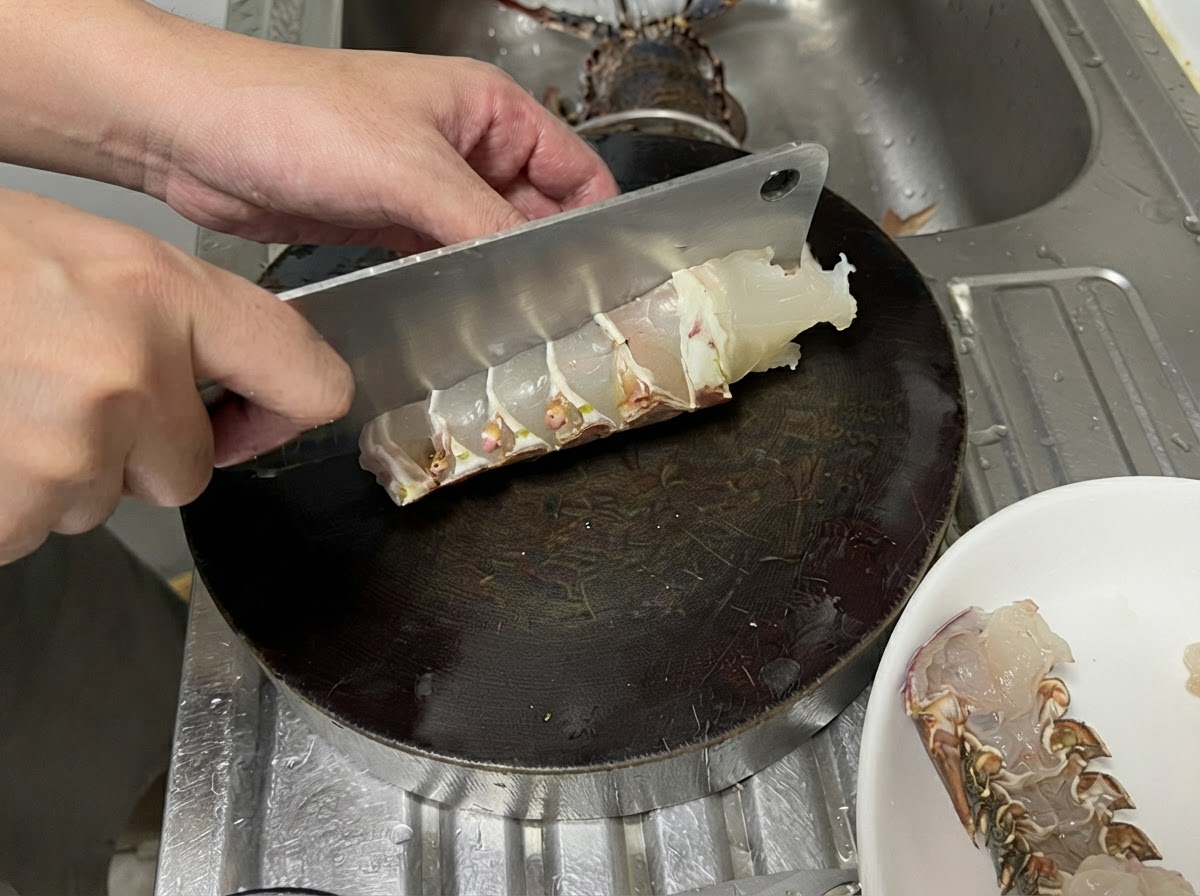 Una grande mannaia di metallo che taglia una coda di aragosta cruda su un tagliere rotondo scuro per creare pezzi spessi e uniformi.