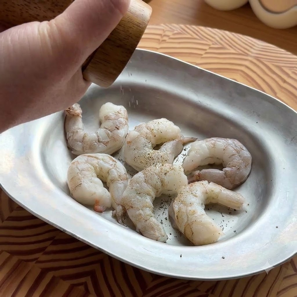 Una mano che utilizza un macinapepe in legno per condire gamberi crudi e sgusciati in un vassoio metallico ovale.
