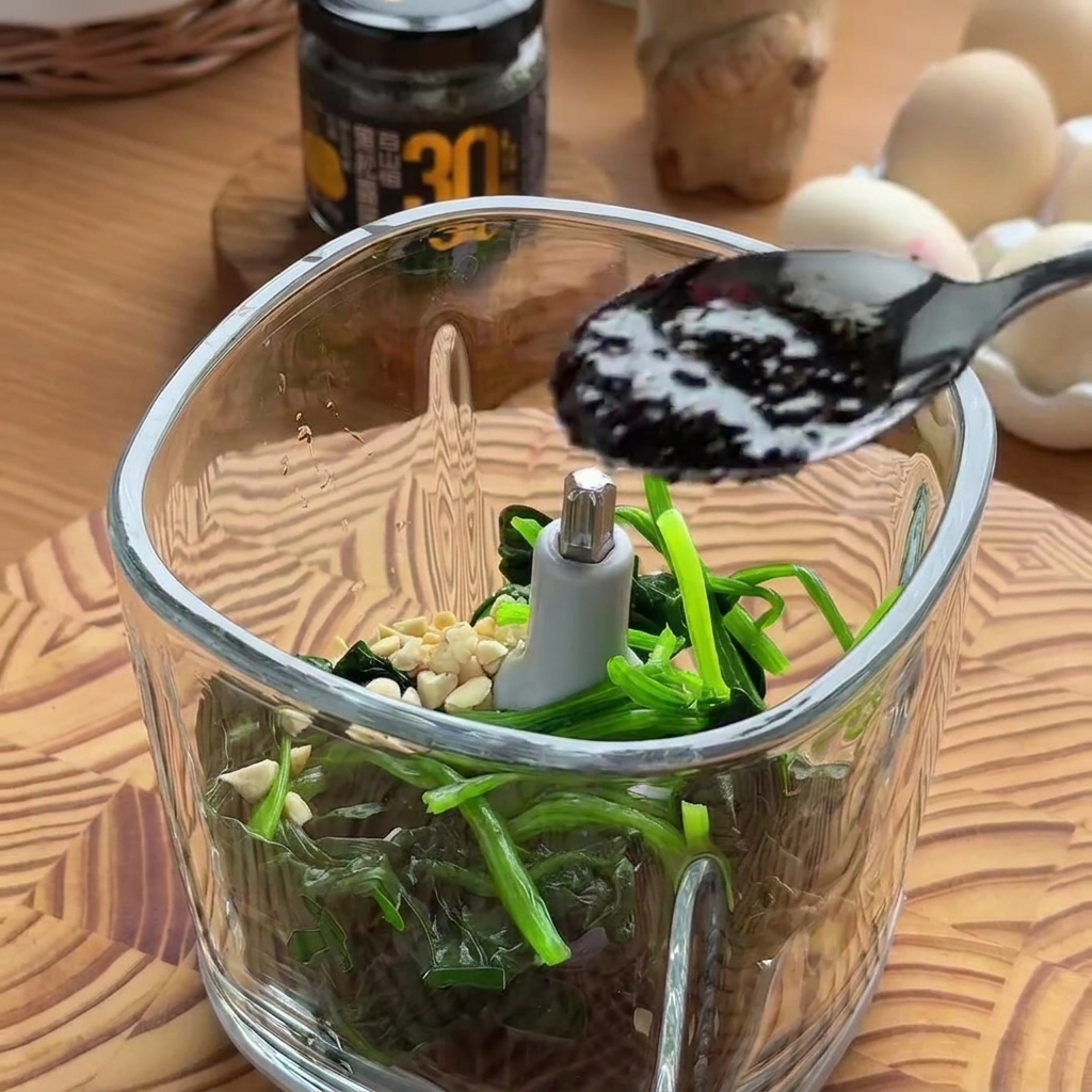 Un cucchiaio nero che versa salsa al tartufo scura e ricca in una ciotola di vetro di un robot da cucina riempita con spinaci e frutta secca.
