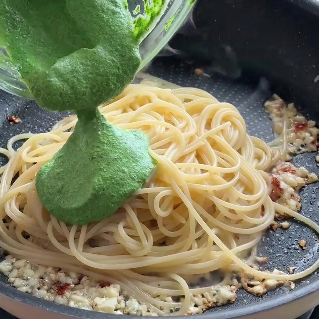 Salsa verde al pesto versata da una ciotola di vetro sugli spaghetti in una padella.