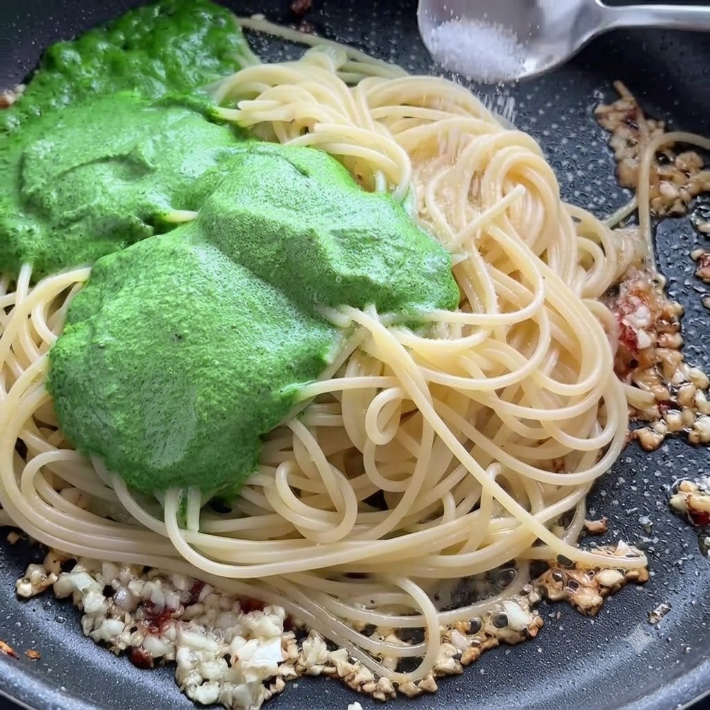 Un cucchiaio di sale cosparso sugli spaghetti e sulla salsa di pesto verde in una padella.
