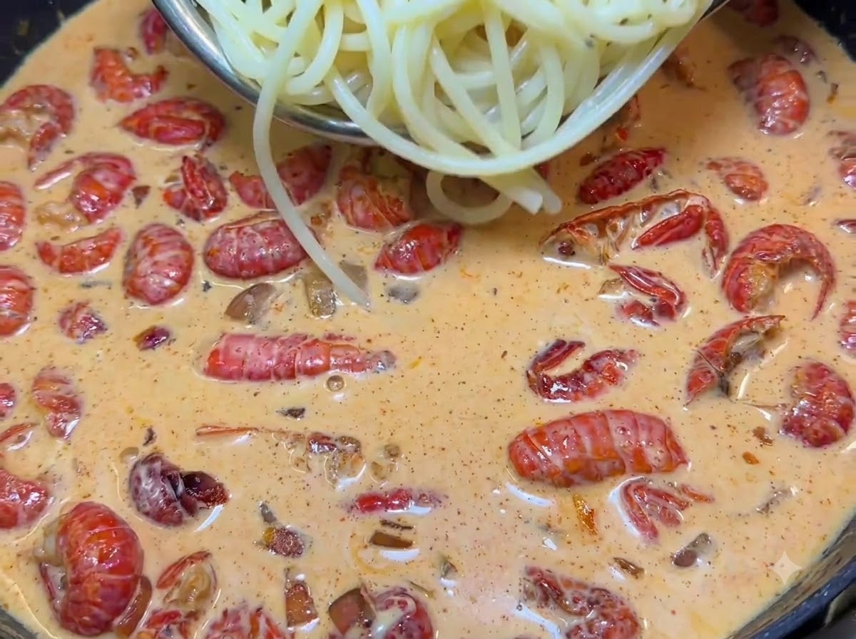 Pinze che sollevano spaghetti bolliti in una padella sfrigolante piena di salsa rosa cremosa e gamberi rossi.