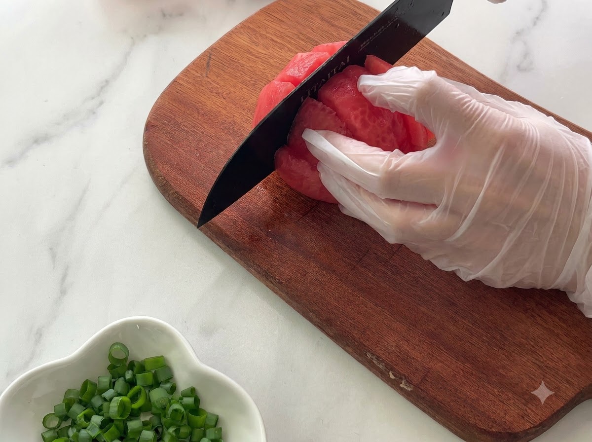 Una mano con i guanti che tiene un pomodoro su un tagliere di legno mentre lo affetta a pezzi con un coltello nero.