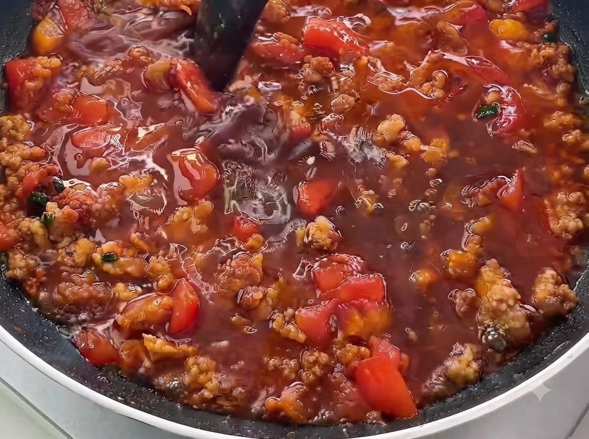 Un primo piano di salsa liquida scura versata in una padella piena di carne macinata dorata e pomodori a cubetti.