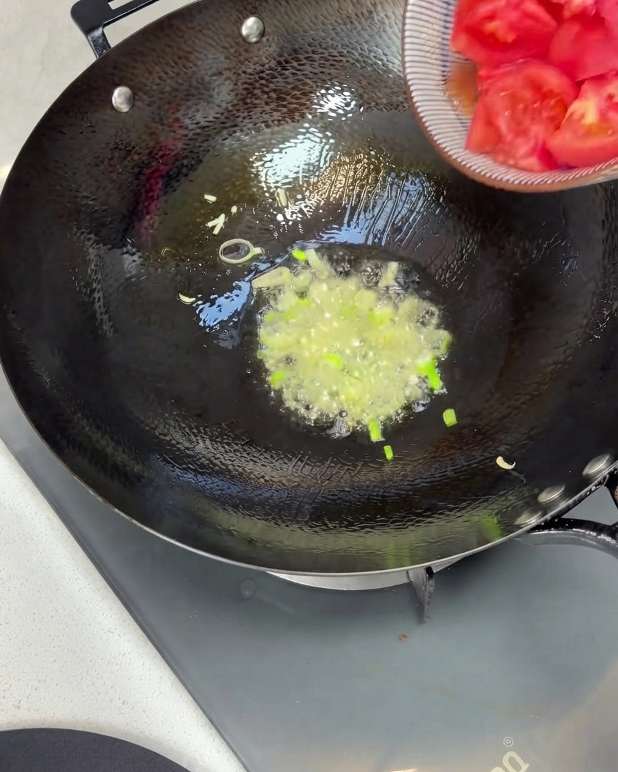 Pezzi di pomodoro trasferiti da una ciotola a un wok con cipollotti saltati e olio.