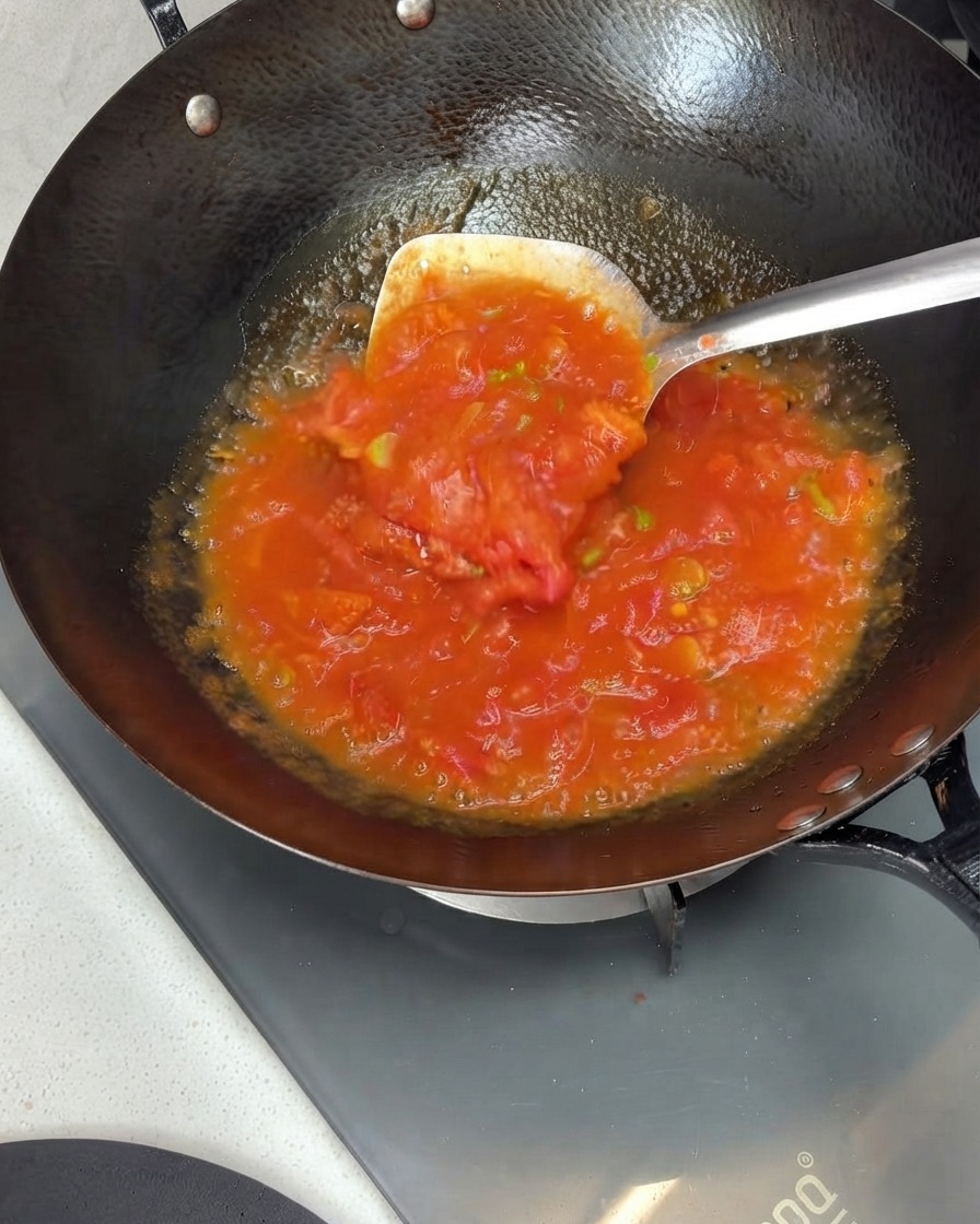 Pomodori schiacciati e mescolati in una ricca salsa allinterno di un wok usando una spatola di metallo.