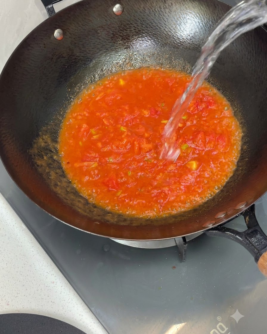 Acqua versata dallalto in un wok pieno di salsa di pomodoro cotta.