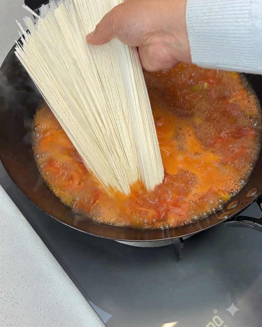 Un mazzetto di noodles secchi tenuto sopra e aggiunto a una zuppa di pomodoro bollente in un wok.