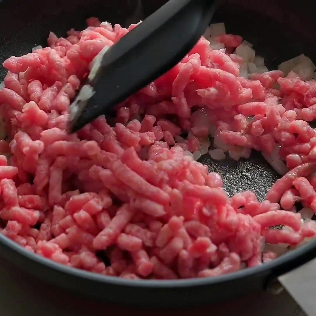 Carne macinata di manzo cruda e rosa sbriciolata e saltata con cipolle in una padella nera.