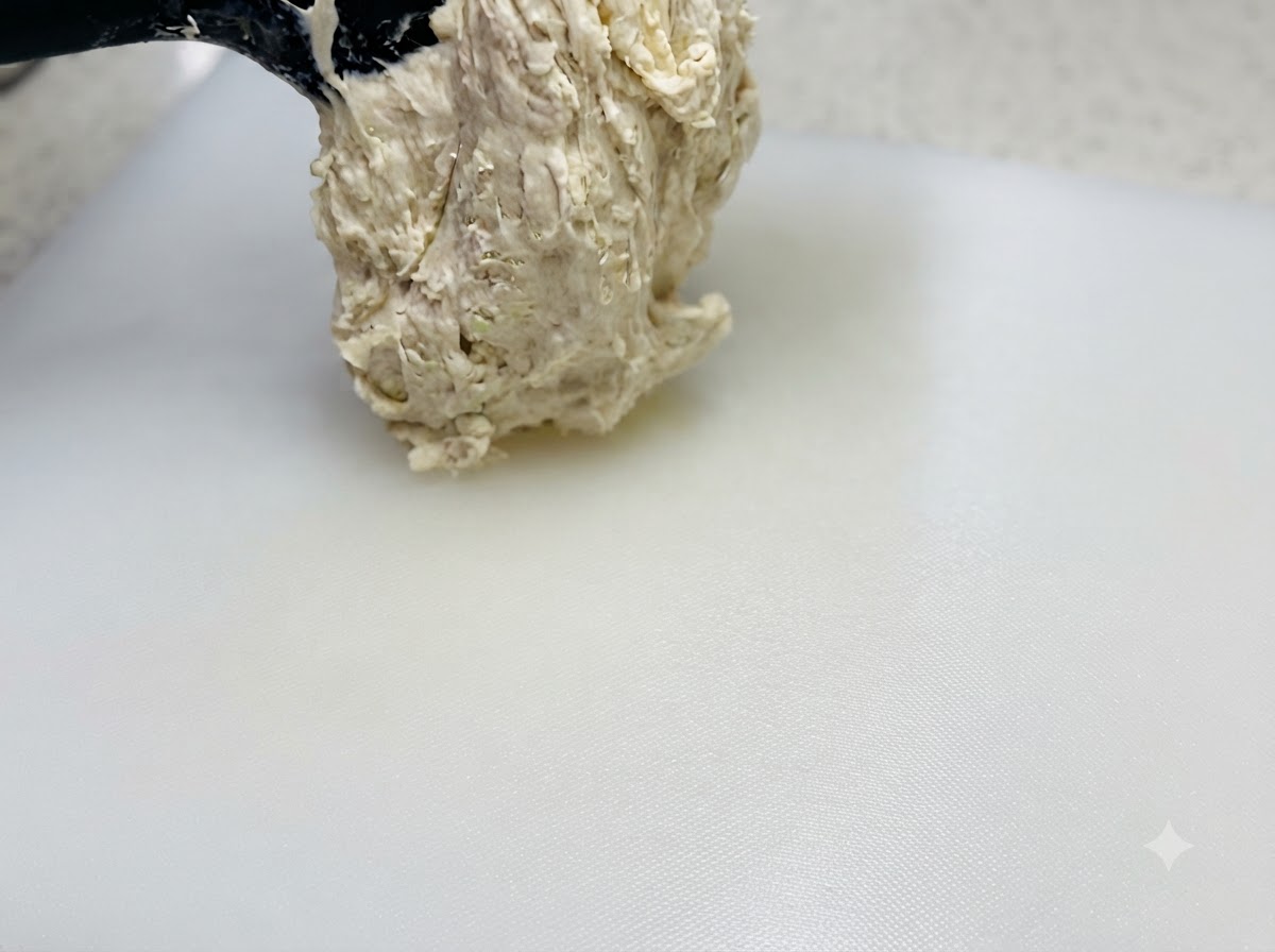 Un impasto appiccicoso ad alta idratazione sollevato con una spatola e posizionato su un tagliere bianco spruzzato dolio.
