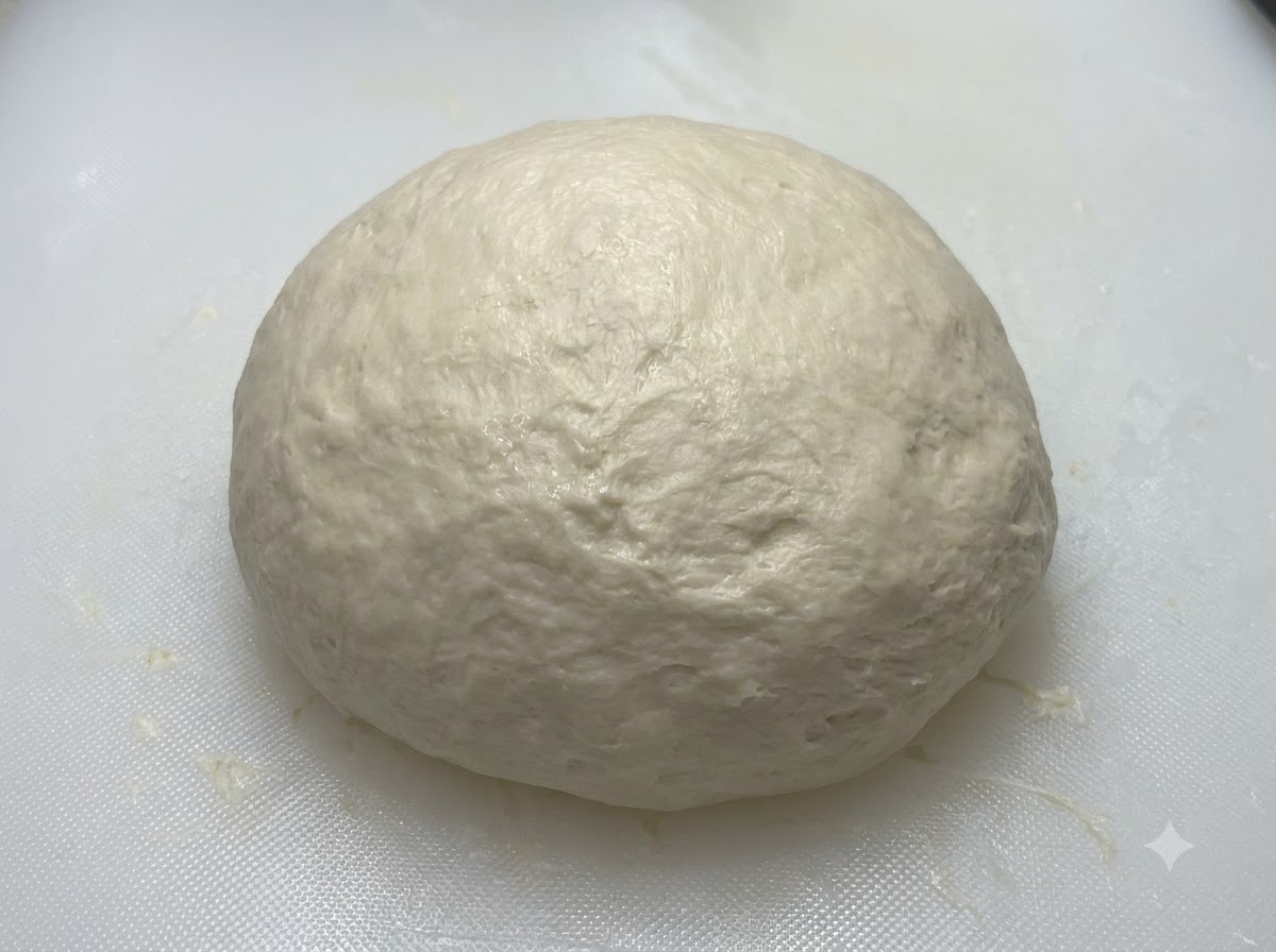 Una pallina di impasto liscia e arrotondata che riposa su un tagliere bianco prima di essere coperta.
