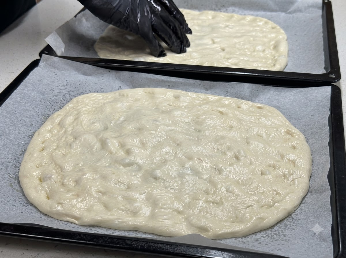 Due teglie con impasto steso e una mano guantata che posiziona metà di pomodorini su tutta la superficie.