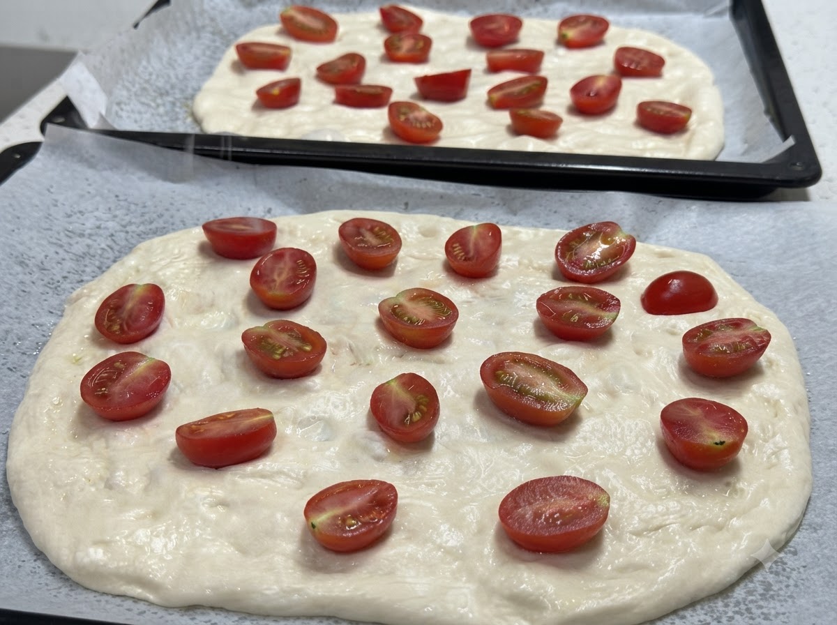 Un primo piano di due basi di pasta per pizza stese e guarnite con pomodorini tagliati a metà, pronte per essere condite.