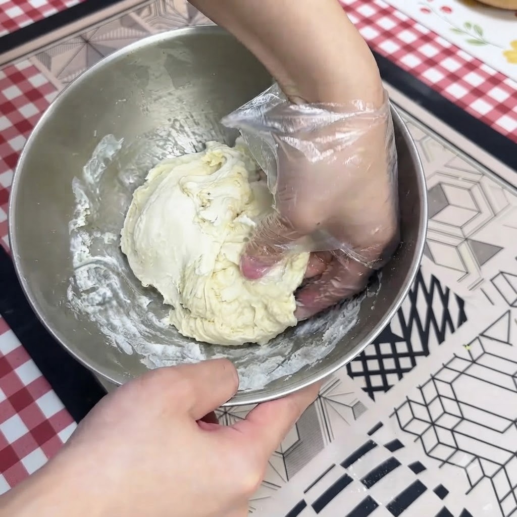 Una mano con guanto che lavora limpasto umido della pizza in una ciotola di acciaio inossidabile.